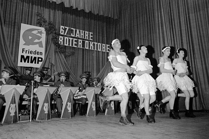 Männer in Frauenkleidern tanzen Ballett, im Hintergrund ein sowjetisches Orchester