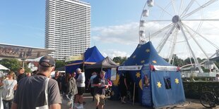 Ein blaues Zelt mit gelben Sternen und der Aufschrift "Wahrsagerin - Lebensberatung mit Herz" auf der Travemünder Woche. Im Hintergrund ein Hochhaus und ein Riesenrad, im Vordergrund Passant*innen.