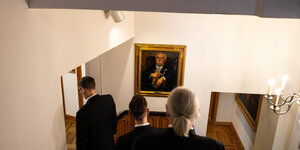 Drei Männer laufen eine Treppe hinunter und dabei auf ein altes Porträtbild zu