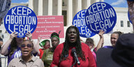 Christian F. Nunes in rotem Kleid, steht an einem Mikrofon und redet. Menschen halten Schilder mit der Aufschrift "Keep Abortion Legal"