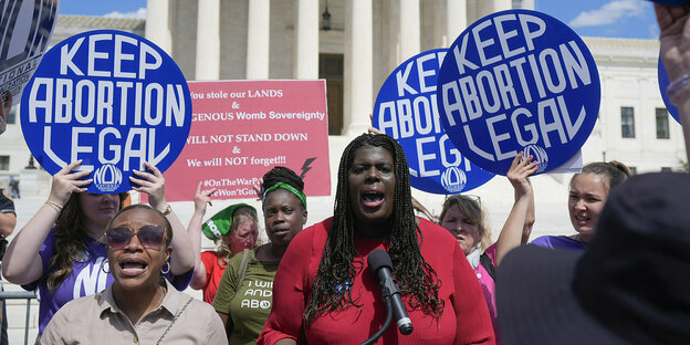 Christian F. Nunes in rotem Kleid, steht an einem Mikrofon und redet. Menschen halten Schilder mit der Aufschrift "Keep Abortion Legal"