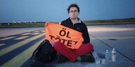 Ein Mann sitzt auf dem Rollfeld eines Flughafens und hält ein Transparent in den Händen, darauf ist zu lesen "Öl tötet"