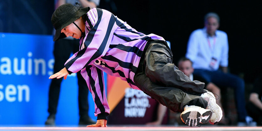 Eine Breakdancerin mit Hut in lila schwarzer Bluse stützt sich seitlich auf Hand und Füße.