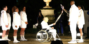 Mehrere Personen in weißer Kleidung in der Nacht. In der Mitte ein älterer Herr im Rollstuhl. Er übergibt das Olympische Feuer an zwei Personen rechts im Bild