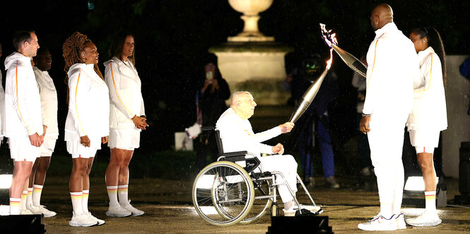 Mehrere Personen in weißer Kleidung in der Nacht. In der Mitte ein älterer Herr im Rollstuhl. Er übergibt das Olympische Feuer an zwei Personen rechts im Bild