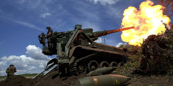 Eine Flamme aus dem Rohr einer Panzerhaubitze. Auf dem Gerät steht ein Soldat, ein anderer steht etwas abseits daneben.