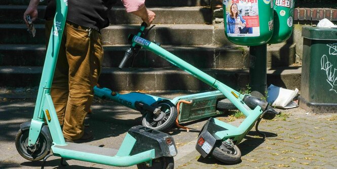 Drei E-Scooter auf einem Gehweg, zwei sind umgefallen. Eine Person versucht einen der beiden wieder aufzurichten.