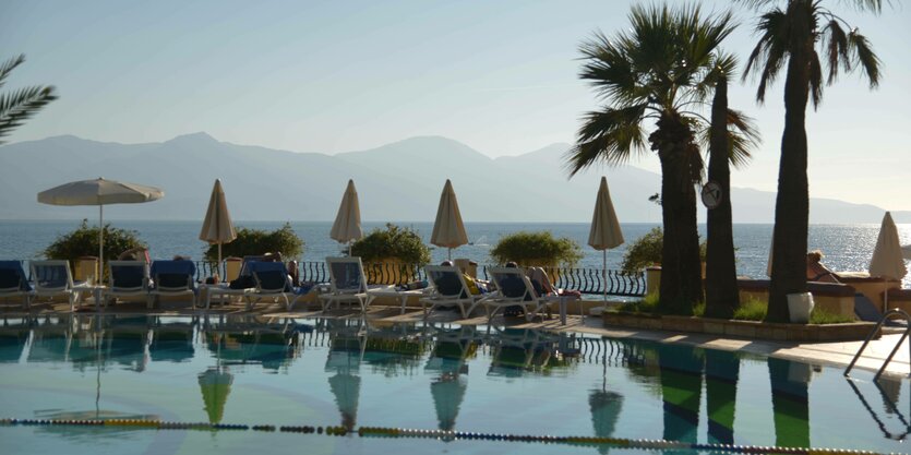 Liegestühle spiegeln sich im Pool, gelbe Sonnenschirme, Meer und eine Küste im Dunst