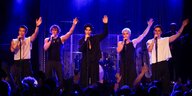 Fünf junge Männer singen auf der Bühne in Mikrofone, synchron heben sie die Hände