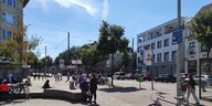 Eine Straßenszene in Bremen-Gröpelingen. Rechts gehen zwei Menschen, links sitzen Menschen auf Steinbänken. Im Hintergrund ein Einkaufszentrum.