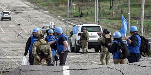 Menschen mit Helmen, teils in Camouflage, teils in blau, laufen eine brüchige Straße entlang.