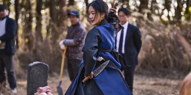 Die Schamanin Lee Hwa-rim, gespielt von Kim Go-eun, steht in einem blauen Gewand und mit Dolchen vor einem Grab und führt ein Ritual zur Geisterbefreiung auf.