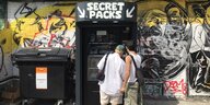 Zwei junge Menschen vor dem Secret Pack Automaten, der aussieht wie ein Snackautomat. Daneben eine große schwarze Mülltonne