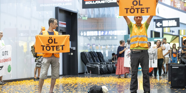 Menschen in orangen Warnwesten, die Schilder mit der Aufschrift "Öl tötet" hochhalten, in einer Flughafenhalle