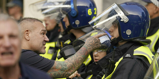 Ein Mann mit tatowirten Armen zeigt auf das Gesicht eines Polizisten, er macht einen sehr wütenden Eindruck