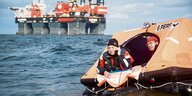 Offenes Meer. Im Vordergrund eine orangefarbene Rettungsinsel, die ein bisschen aussieht wie ein schwimmendes Zelt. Darin sitzen zwei Menschen in rot-schwarzen Anzügen. Eine von beiden hat ein klobiges Mobiltelefon in der Hand. Im Hintergrund ein riesiges Kranschiff.