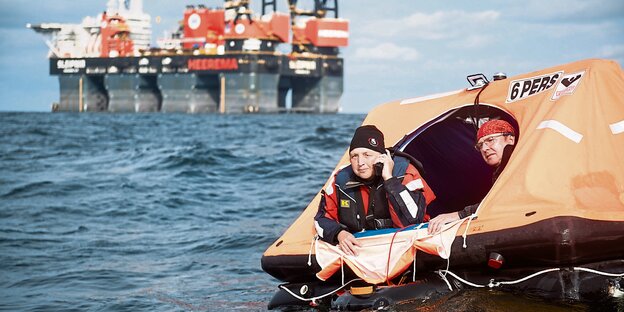 Offenes Meer. Im Vordergrund eine orangefarbene Rettungsinsel, die ein bisschen aussieht wie ein schwimmendes Zelt. Darin sitzen zwei Menschen in rot-schwarzen Anzügen. Eine von beiden hat ein klobiges Mobiltelefon in der Hand. Im Hintergrund ein riesiges