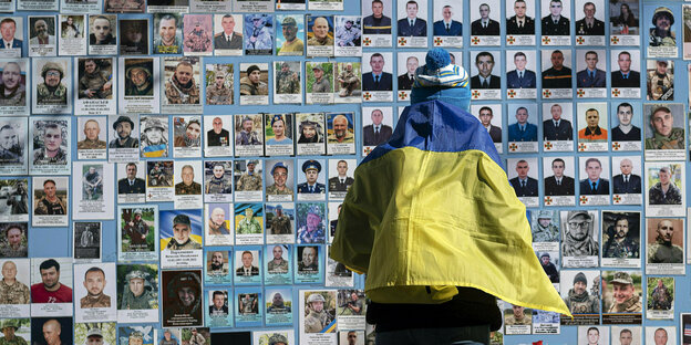 Eine in eine ukrainische Flagge gehüllte Frau steht neben der Gedenkmauer für die gefallenen Soldaten der Ukraine. Zu sehen sind viele kleine Fotos mit den Namen und dem Todestag der toten Soldaten