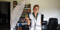 Eine ältere kurzhaarige Frau hat in einer Wohnung einen Schirm aufgespannt, sie trägt eine weiße Warnwest und einen anhänger "Omas gegen rechts"