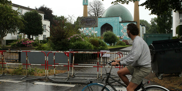 Die Blaue Moschee, eigentlich Imam-Ali-Moschee, in Hamburg nach dem Verbot des Islamischen Zentrum Hamburg (IZH). Ein vorbeifahrender Fahrradfahrer schaut zu dem Gebäude hinter einer Absperrung aus rot-weißen Gittern