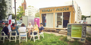 Der mobile Nachbarschaftstreff mit Namen Kleiner Frieder ist ein kleines Holzhaus auf Rädern - es sitzen Menschen in einem Stuhlkreis davor und tauschen sich aus