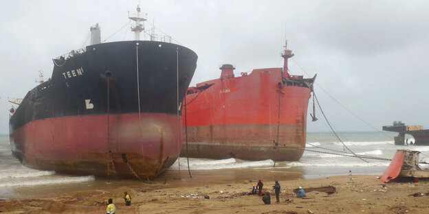 Zwei große Schiffe liegen an einem Strand, davor laufen Arbeiter herum