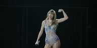 Taylor Swift steht in einem paillettenbesetzten Bodysuit auf der Bühne und spannt als Geste den Bizeps an.