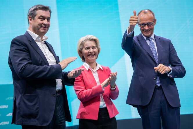 Markus Söder, Ursular von der Leyen und Friedrich Merz stehen auf einer Wahlkampfbühne, Söder und von der Leyen beklatschen etwas, Merz macht eine Daumen-Hoch-Geste