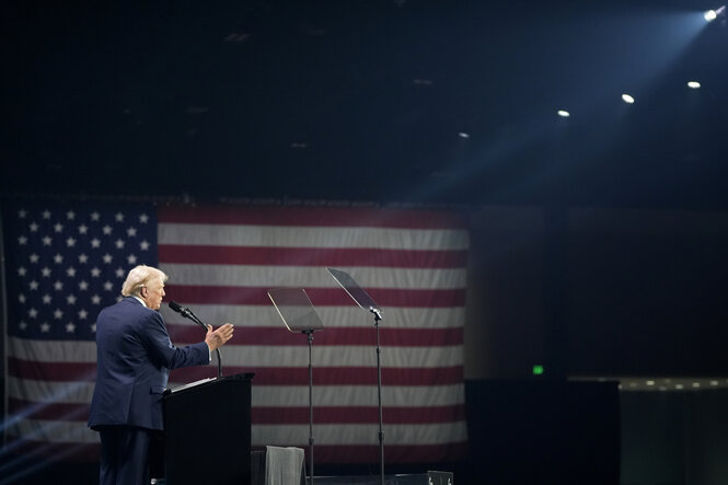 Aus der Distanz fotografiert, steht Donald Trump allein auf einer Bühne vor einem Rednerpult und spricht, ein Scheinwerfer beleuchtet ihn, im Hintergrund hängt eine große US-Flagge