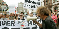 Vor einer Gruppe von Demonstrant*innen mit Transparenten und Plakaten spricht ein Mann in ein Megafon