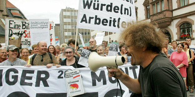 Vor einer Gruppe von Demonstrant*innen mit Transparenten und Plakaten spricht ein Mann in ein Megafon