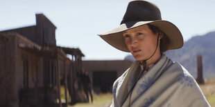 Vivienne Le Coudy, gespielt von Vicky Krieps, steht in Tunika und einem Cowboy-Hut vor einem Cottage im Western-Stil. Im Hintergrund sind Berge.