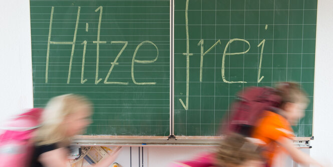 Grundschüler mit Schulranzen rennen an einer grünen Tafel vorbei, auf der mit Kreide "Hitzefrei" geschrieben steht.