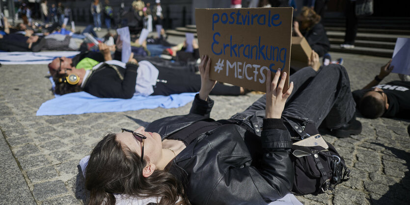 Eine Frau liegt auf gepflastertem Boden vor dem Roten Rathaus in Berlin. Sie hält ein Schild mit der Aufschrift "Postvirale Erkrankung #ME/CFS" in der Hand