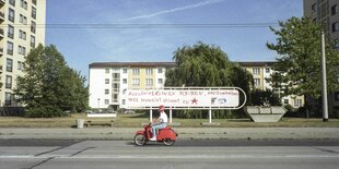 Person auf einem roten Roller mit einem roten Helm fährt an Wohnhäusern vorbei. Auf ein langes Straßenschild im Hintergrund steht in krakeliger, großer roter Schrift: "Ausländerinnen bleiben, Nazis vertreiben, wer schweigt stimmt zu"