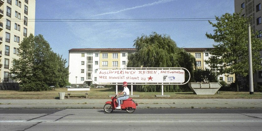 Person auf einem roten Roller mit einem roten Helm fährt an Wohnhäusern vorbei. Auf ein langes Straßenschild im Hintergrund steht in krakeliger, großer roter Schrift: "Ausländerinnen bleiben, Nazis vertreiben, wer schweigt stimmt zu"