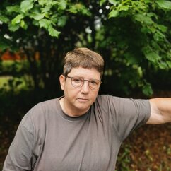 Sylvia Rademacher trägt ein graues T-Shirt und hat kurze Haare und Brille