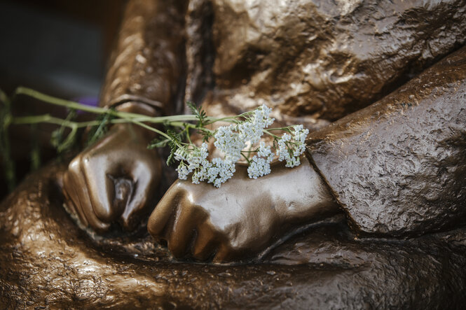 Hände eine Bronzestatue, Blüten der Schafgarbe sind darauf abgelegt