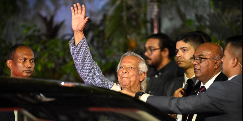 Muhammad Yunus hat längere graue Haare, hebt die Hand und winkt