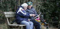 Zwei ältere Männer lesen auf einer Parkbank sitzend die Zeitung.