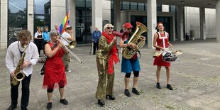 Fünf Personen in bunten Kostümen mit Blasinstrumenten, Schlagzeug und Megafon stehen vor einem grauen Haus, dem Rathaus Mitte