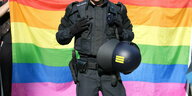 Der Rumpf eines Polizeibeamten in schwarzer Montur, einen Helm in der Hand, ist vor einer Regenbogenflagge zu sehen