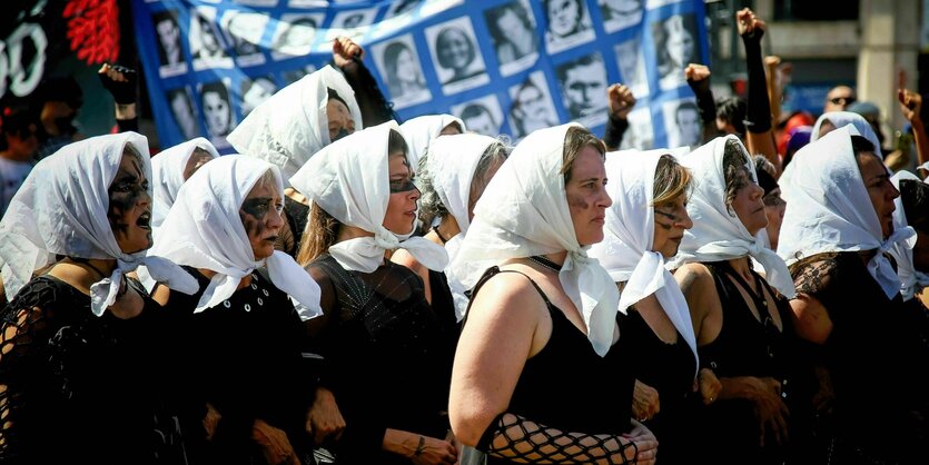 Demonstranten tragen weiße Schals auf dem Kopf während einer Demonstration, im Hintergrund ein Banner mit Portraits von Opfern der Diktatur