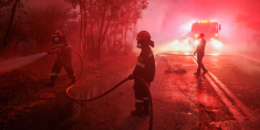 Drei Feuerwehrleute stehen im Dunkeln auf einer Waldstraße. Ein Feuerwehrmann versucht etwas, was nicht im Bild ist, zu löschen. Im Hintergrund dichte Rauchschwaden und die Scheinwerfer eines Feuerwehrautos.