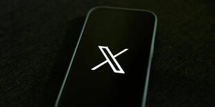 Das Logo von X auf der Rückseite eines stilisierten Smartphones vor schwarzem Hintergrund