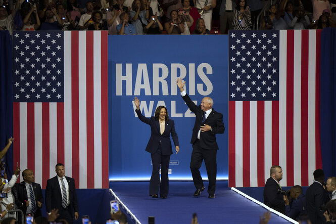 Aus der Distanz gesehen: Kamala Harris und Tim Walz stehen auf einer Wahlkampfbühne und winken dem Publikum vor ihnen zu