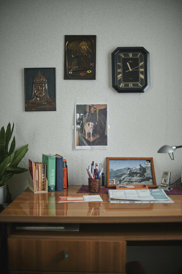 Ein Holzschreibtisch, auf dem Bücher stehen, an der Wand hängen Bilder und eine Uhr
