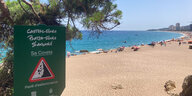 Blick auf einen Strand mit Sonnenbadenden, im Vordergrund ein Schild, das vor Steinschlag warnt