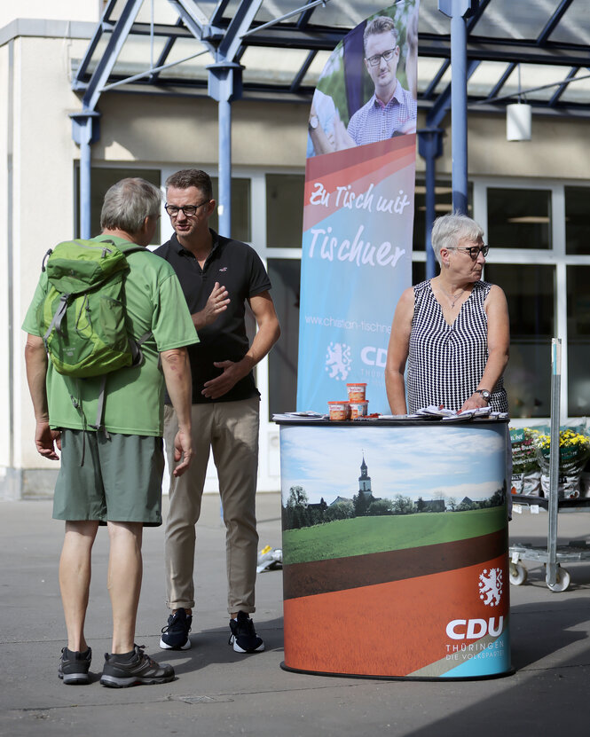 Vor einem Plakat mit der Aufschrift "Zu Tisch mit Tischner" stehen drei Personen. Ein Mann im schwarzen Ti-Shirt diskutiert mit einem mann in grüner Kleidung und mit grünem Rucksack. Eine Frau mit kurzen grauen Haaren steht daneben und schaut von den beiden Männern weg