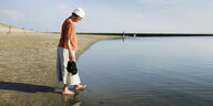 Eine Person mit Sonnenmütze steht am Strand und befühlt das Wasser des Meeres mit einem ihrer Füße